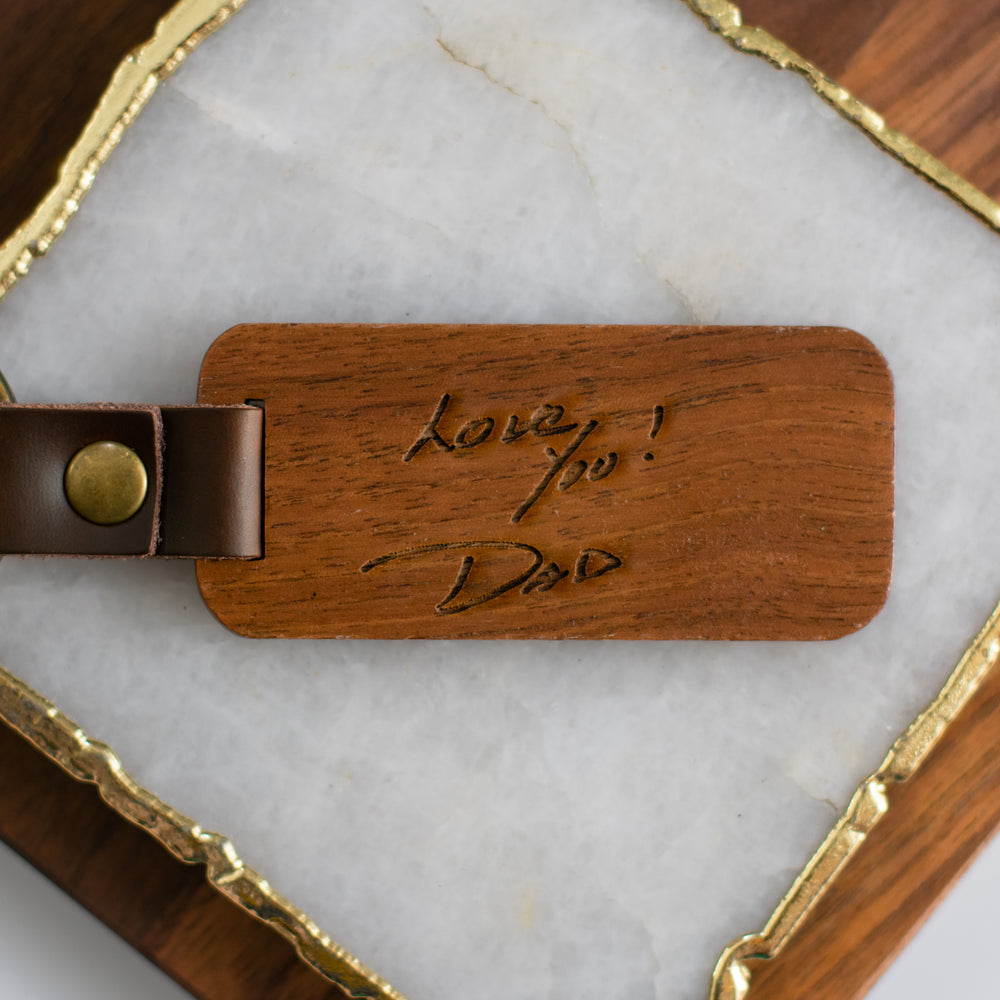 A close-up of a wooden keychain with Love You! Dad engraved on it, sitting in the middle of a gold-framed granite surface.