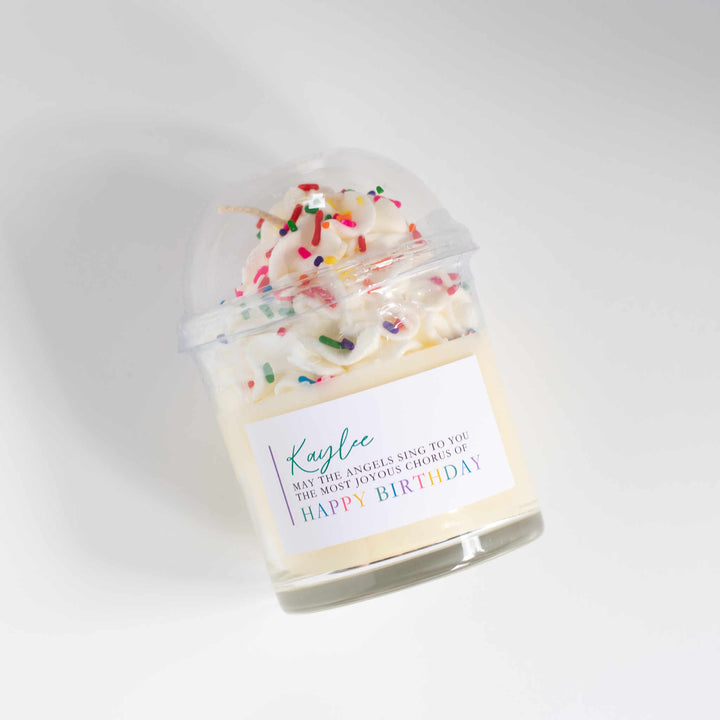 A glass candle labeled Kaylee May the angels sing to you the most joyous chorus of Happy Birthday with whipped cream and sparkles at the top with a plastic lid. Tilted to the left on a white background.