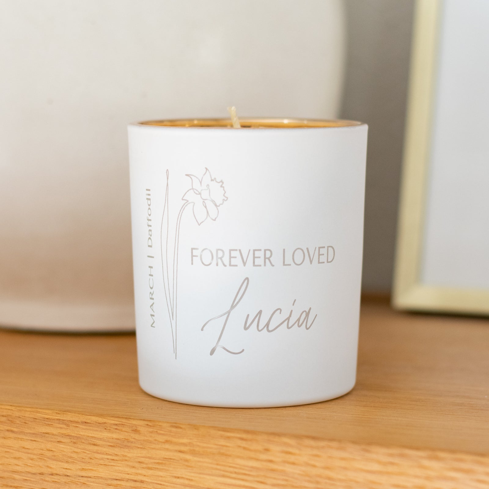 A white-coated candle with engraved text Forever Loved Lucia and a flower on the left. Sitting on a wooden surface.