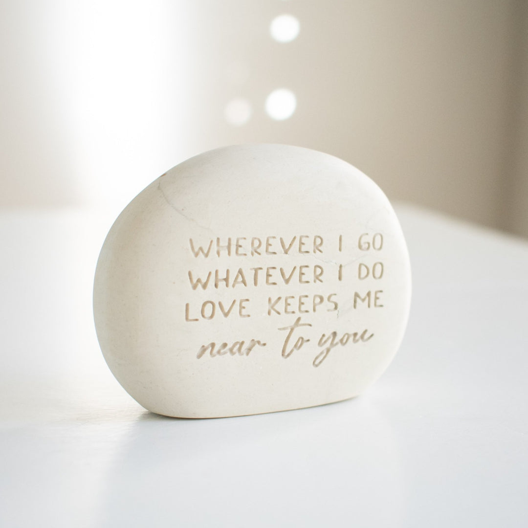 A memorial gemstone with Whenever I Go Whatever I Do Love Keeps Me Near To You engraved on it sitting outside on a white surface angled to the right