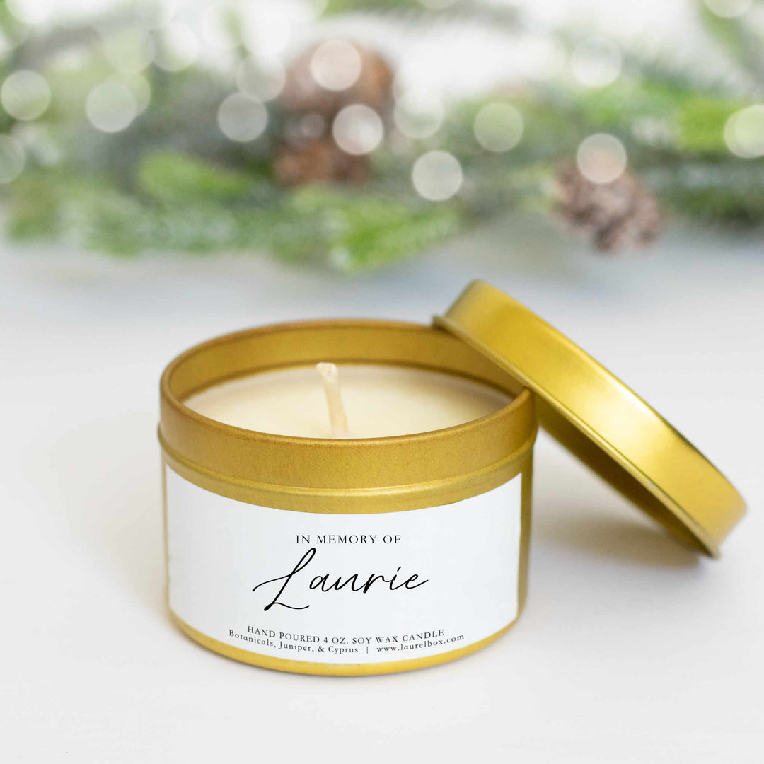 A gold-coated wide candle with a white sticker with the text in Memory of Laurie sitting on a white table