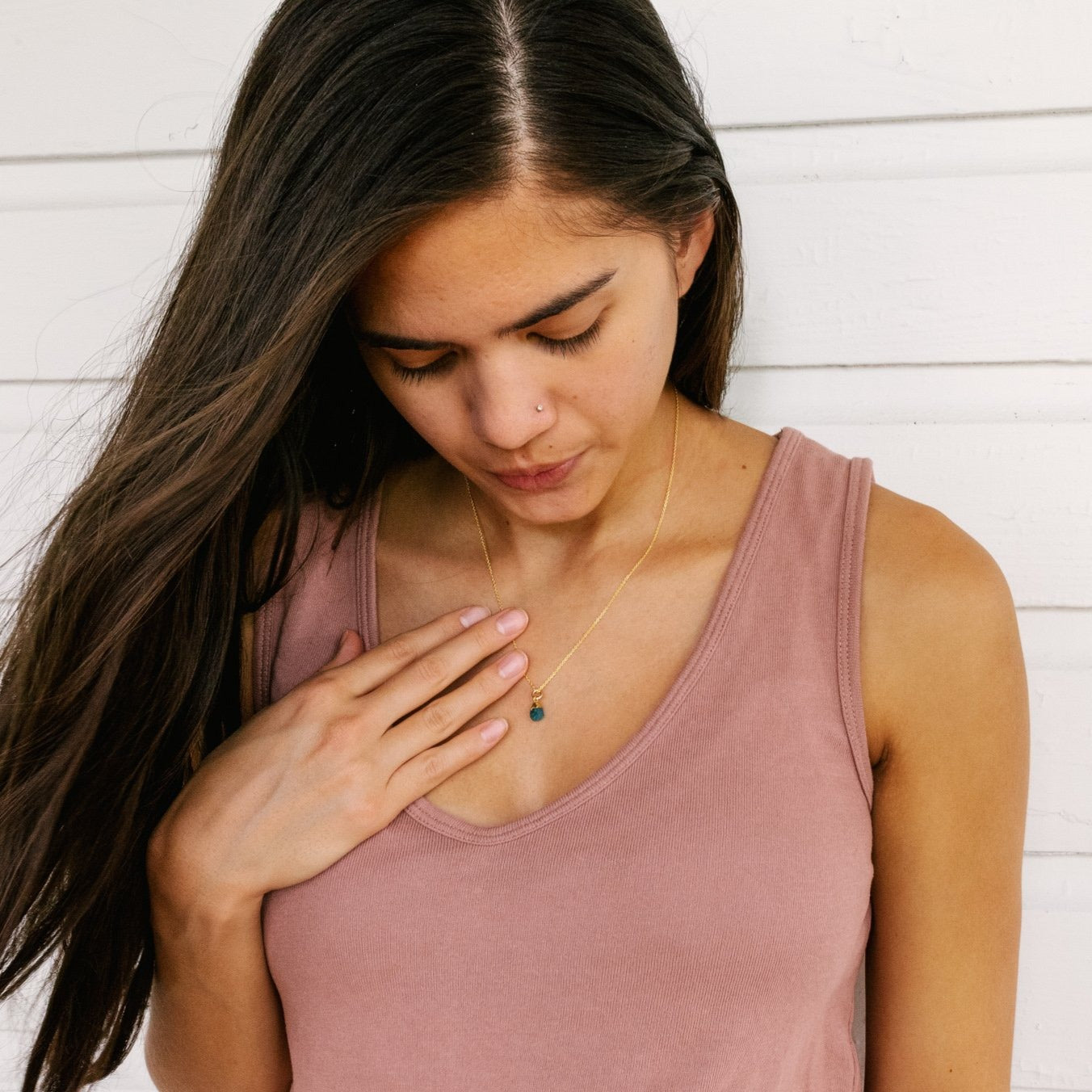 photo of a woman with long brown hair wearing a dark pink tank top - holding her hand to her chest next to a green gemstone necklace on a gold chain