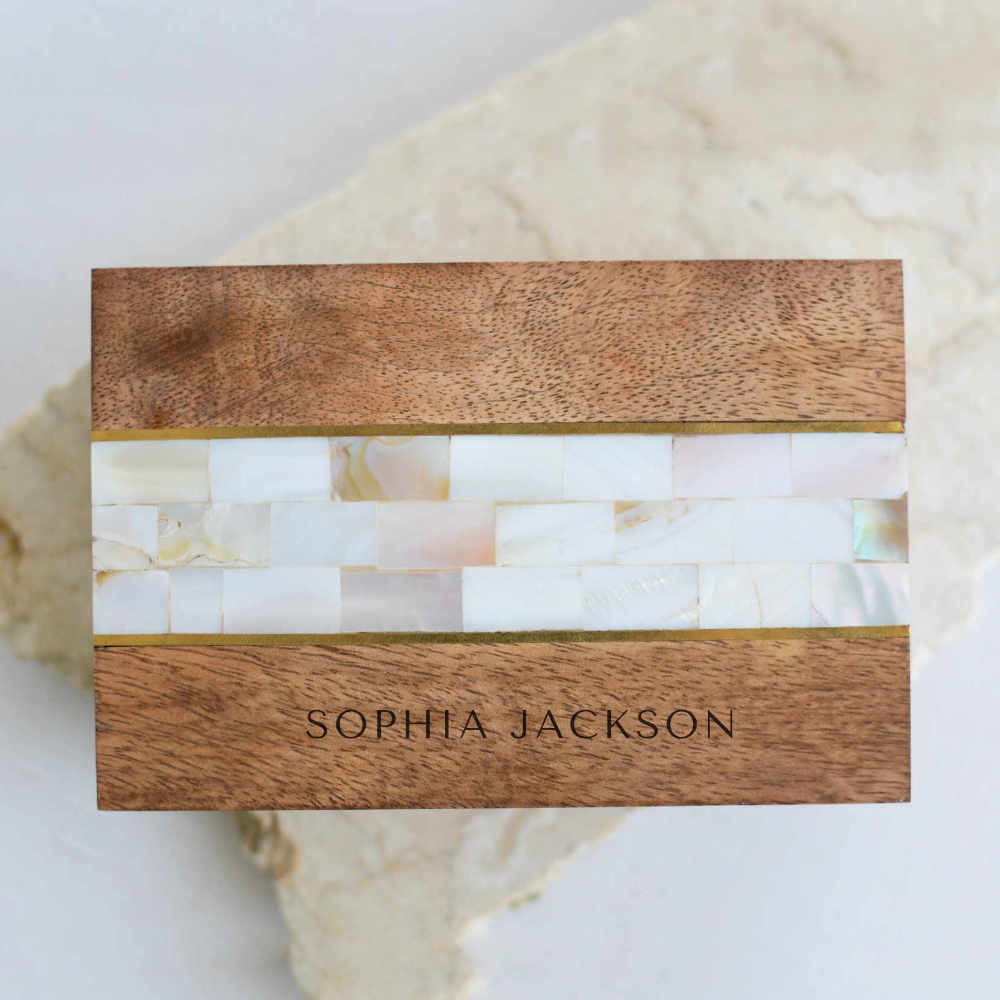 A close-up of a wooden memory box with mother-of-pearl stones in the middle, with an engraved Sophia Jackson text at the bottom, and placed on top of a granite surface.