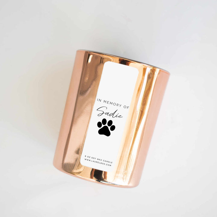 A close-up of a gold candle with a white label with the text "In Memory of Sadie" with a paw print against a white background.