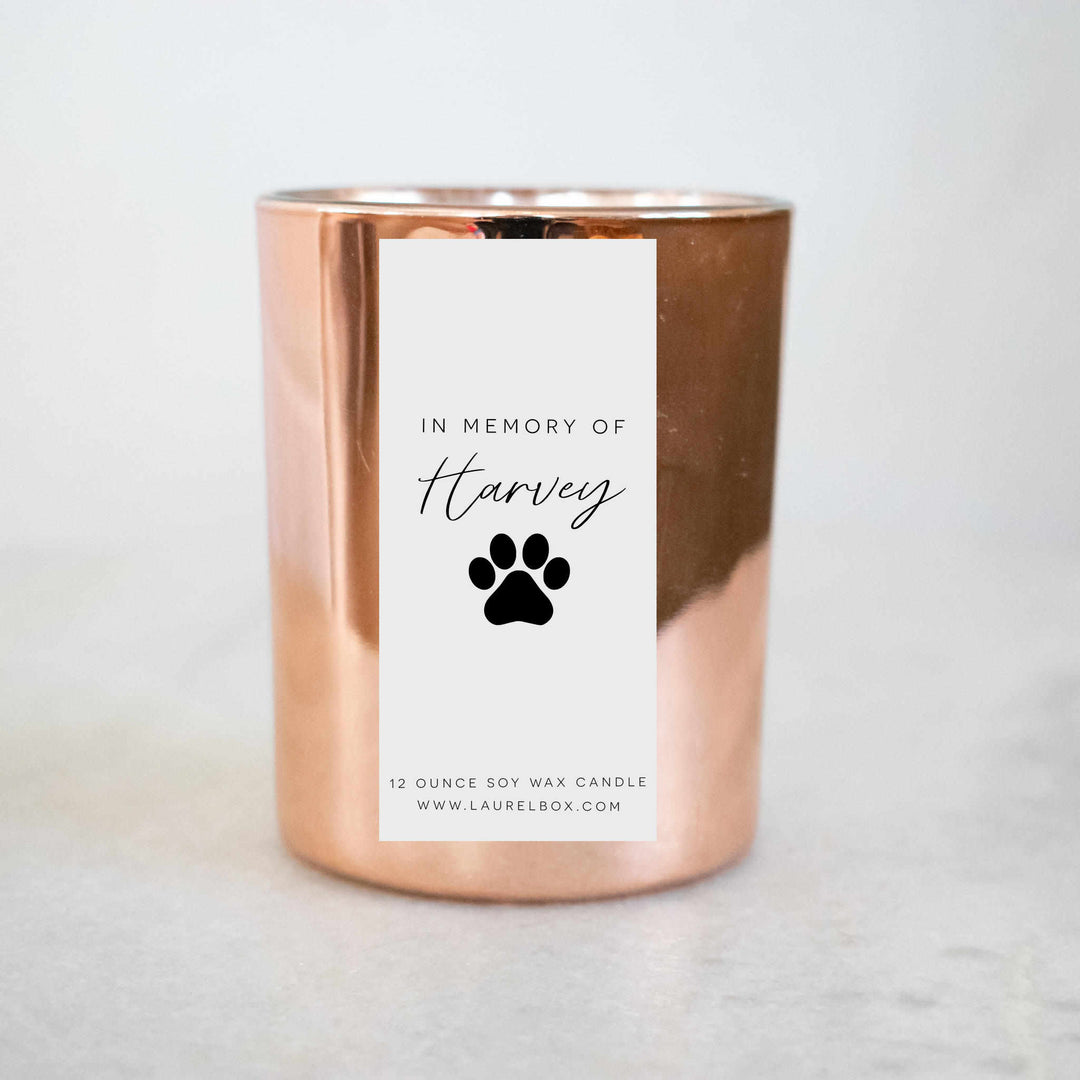 A close-up of a gold candle with a white label with the text "In Memory of Harvey" with a paw print against a white background.