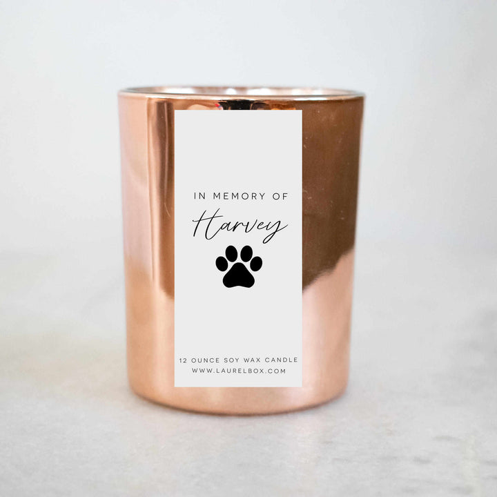 A close-up of a gold candle with a white label with the text "In Memory of Harvey" with a paw print against a white background.