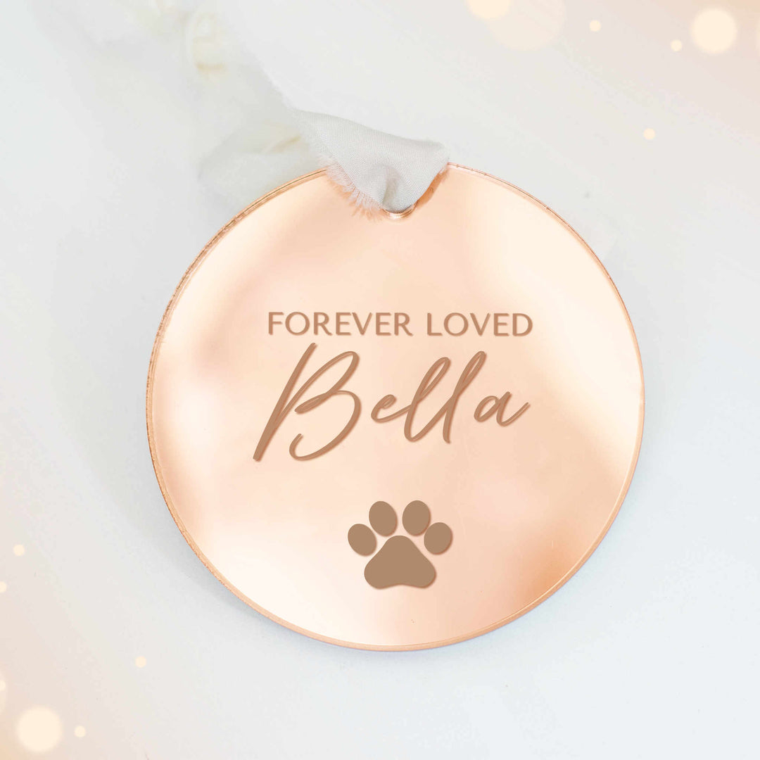 A personalized gold mirrored ornament featuring the text "Forever Loved" and "Bella", with a paw print symbol engraved, and a white ribbon at the top, set against a white background.