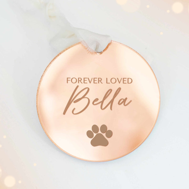 A personalized gold mirrored ornament featuring the text "Forever Loved" and "Bella", with a paw print symbol engraved, and a white ribbon at the top, set against a white background.