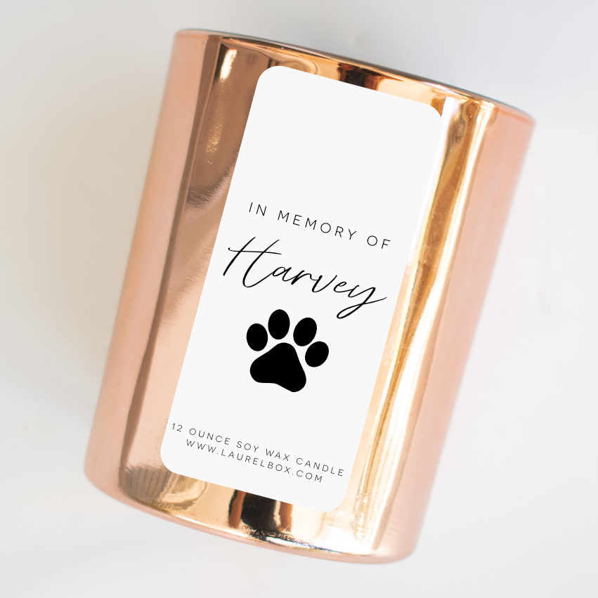 A gold-coated candle with a white sticker, the text in Memory of Harvey, and a paw print. Tilted to the right against a white background