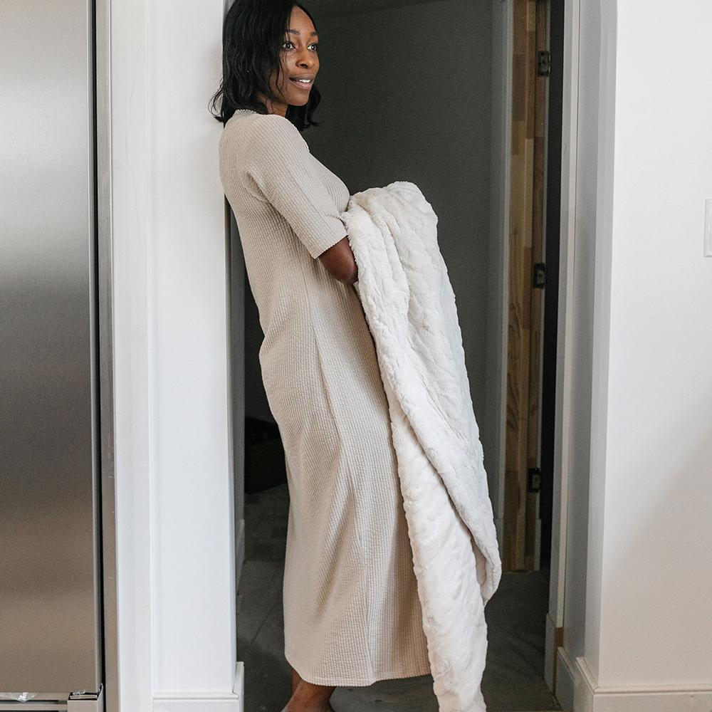 A woman wearing a long striped dress stands against a white wall, holding onto a fuzzy white blanket.