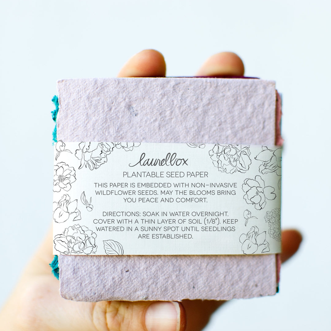 photo of a hand holding pastel square pieces of paper in multiple colors stacked on top of one and other with a with label holding them all together that displays"laurelbox Plantable Seed Paper"