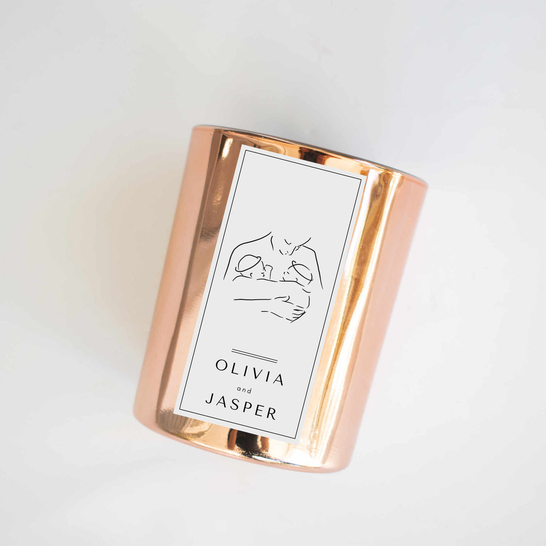 A gold-coated candle with a white sticker, the text Olivia and Jasper, and a woman holding two twins. Tilted to the right against a white background