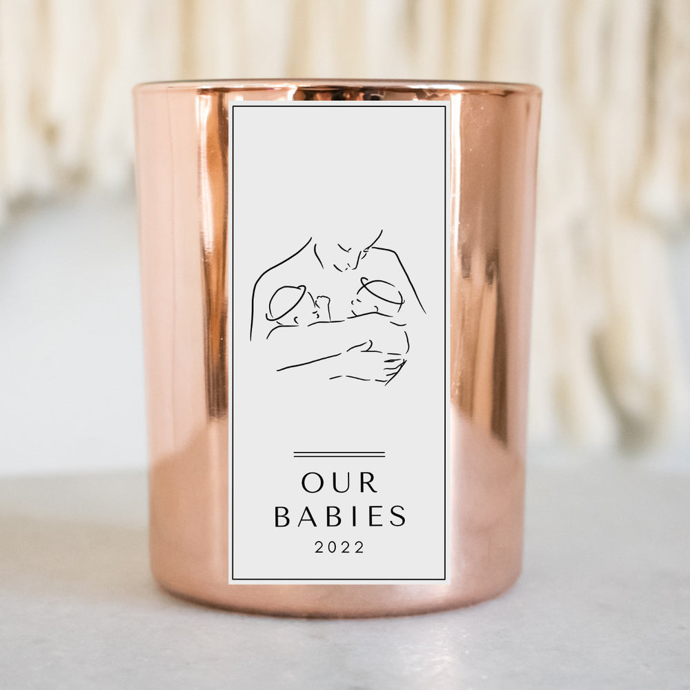 A gold-coated candle with a white sticker, the text Olivia and Jasper, and a woman holding two twins sitting on a white surface