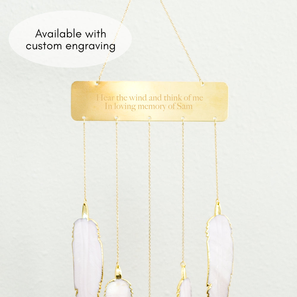 A wind chime hanging against a white wall features five mother-of-pearl-shaped feathers with a rectangular bar at the top with a text engraved hear the wind and think of me in loving memory of Sam