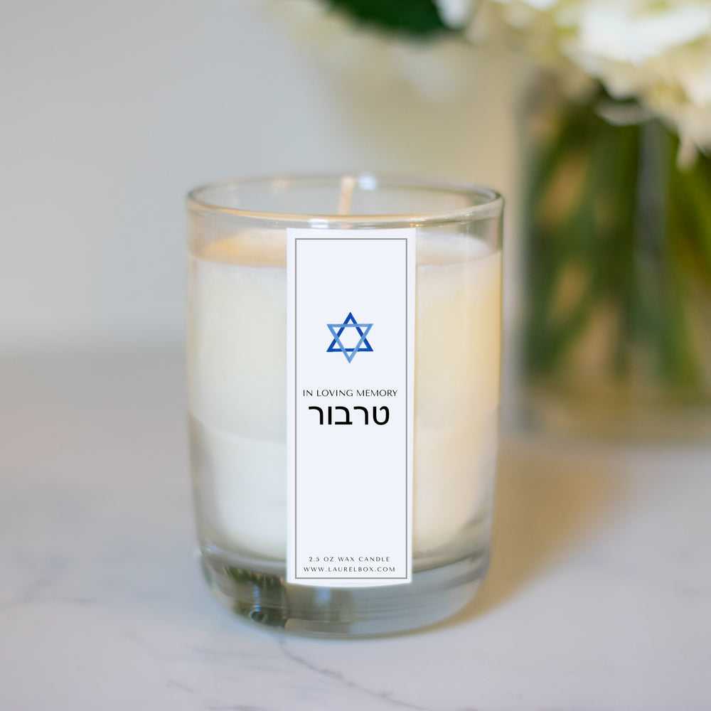 A glass candle with a white sticker sitting on a white surface with a vase of flowers in the background