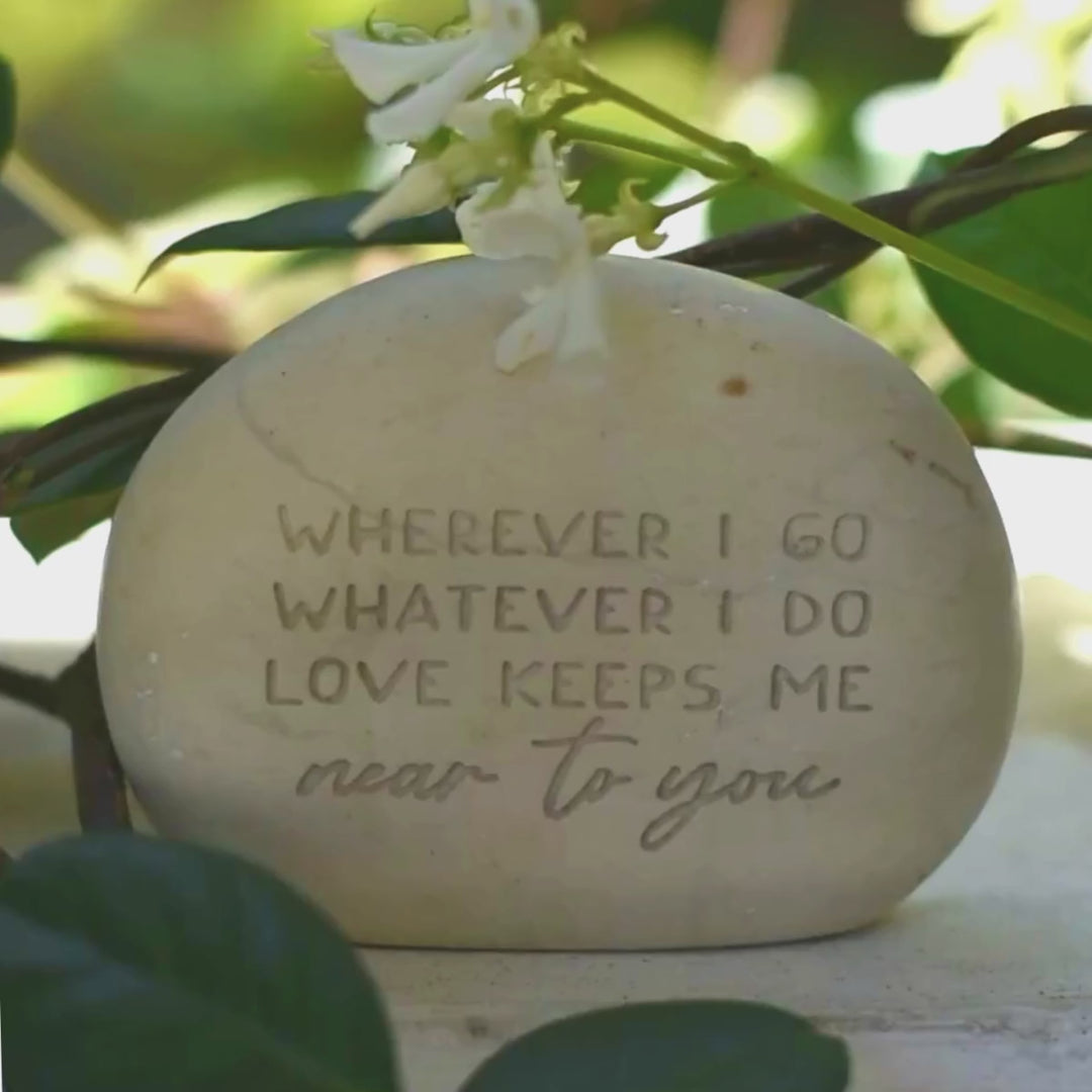A memorial gemstone with Whenever I Go Whatever I Do Love Keeps Me Near To You engraved on it sitting outside next to flowers