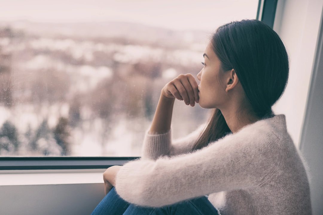 A young woman with long dark hair sits by a window, gazing outside with a pensive expression, wearing a light sweater.