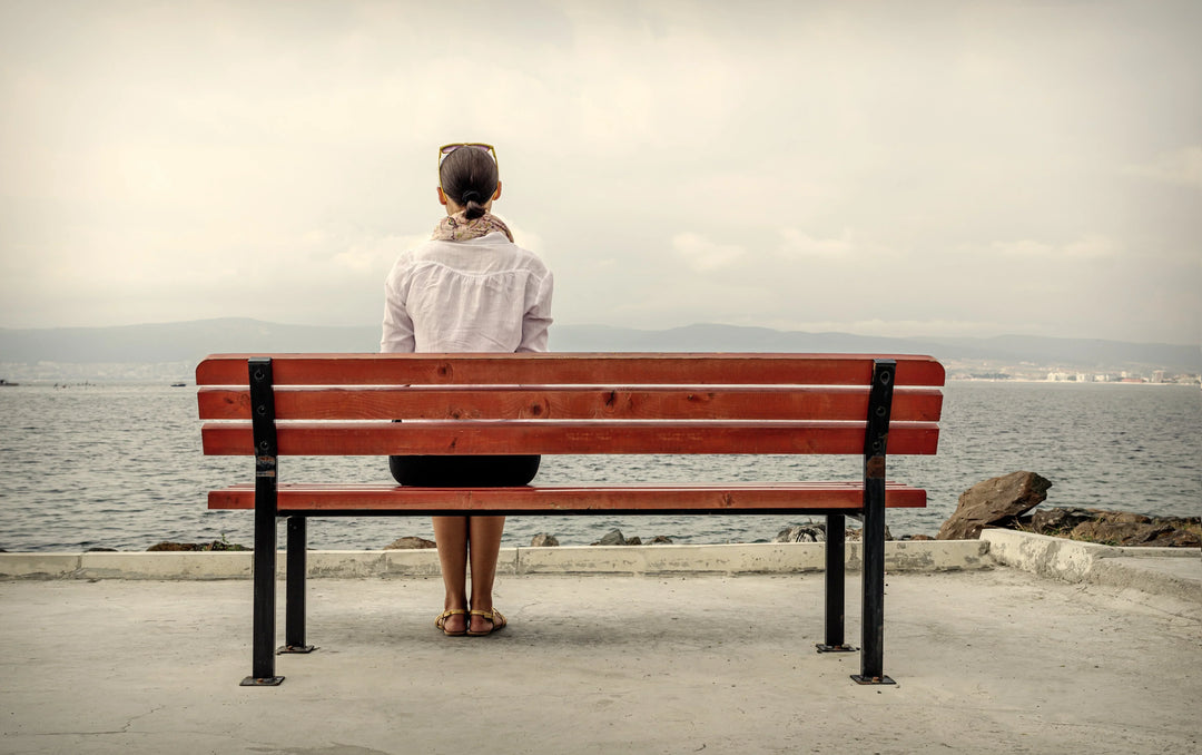 A woman in a light blouse sits on a wooden bench facing the ocean, reflecting on her thoughts by the water.