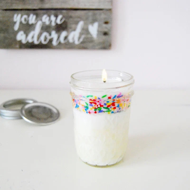 Candle in a glass jar with white wax and colorful floral decoration, lit, alongside metal lids on a white surface.