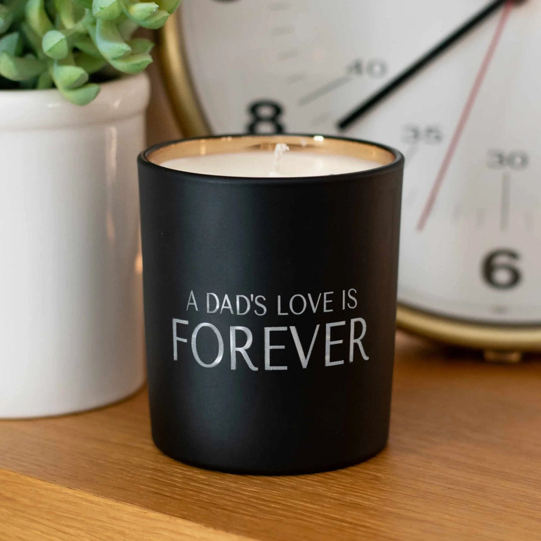Black candle with gold trim, engraved with 'A DAD'S LOVE IS FOREVER,' placed beside a white pot and clock. 15 Heartfelt Sympathy Gifts for Losing a Dad, Father’s Day 2026 Edition.