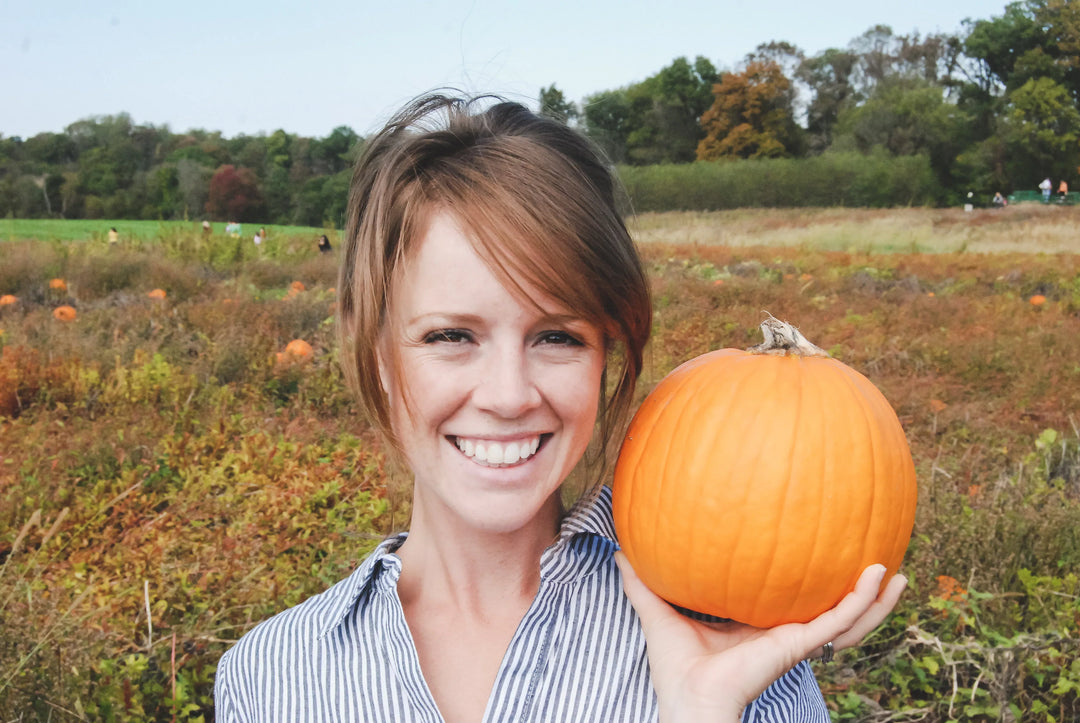 Woman with short hair smiling, holding a bright orange pumpkin in a pumpkin patch during autumn.