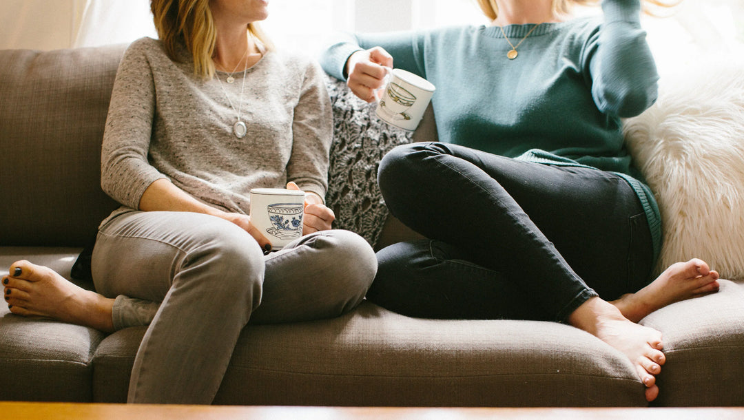 Two women sitting on a couch, holding mugs, wearing casual sweaters and jeans, in a cozy indoor setting.