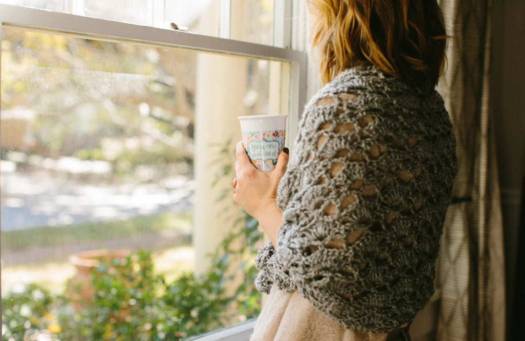 A person with wavy hair wearing a gray crochet shawl while holding a cup, looking out a window at greenery.