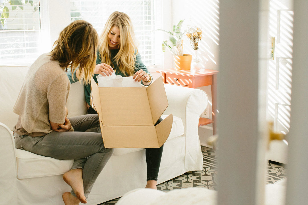 Two women opening a cardboard gift box with tissue paper on a white sofa in a bright room, showing a thoughtful moment.
