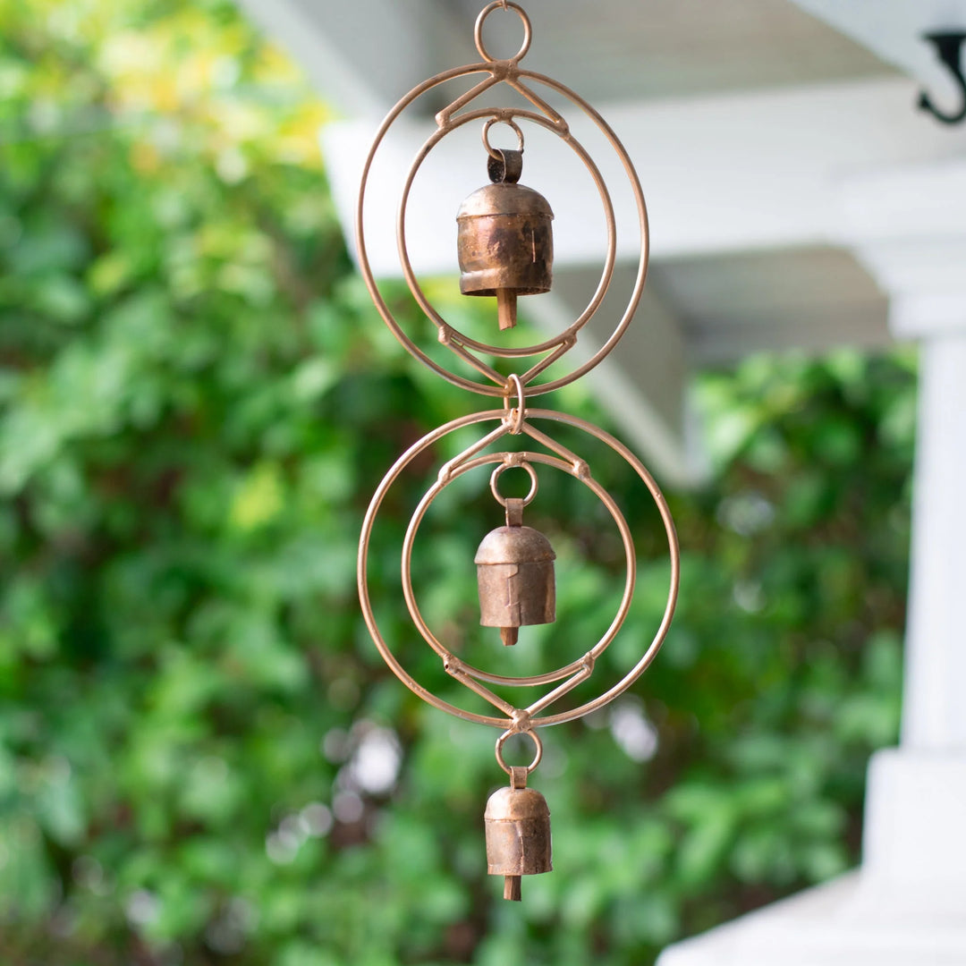 Wind chime with three bronze bells on circular metal rings, hanging outdoors with greenery in the background. Healing Grief Through Sound: The Power of Wind Chimes.