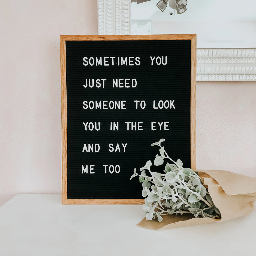 Black letter board in wooden frame with white text saying 'Sometimes you just need someone to look you in the eye and say me too'. Green plant in brown paper beside.
