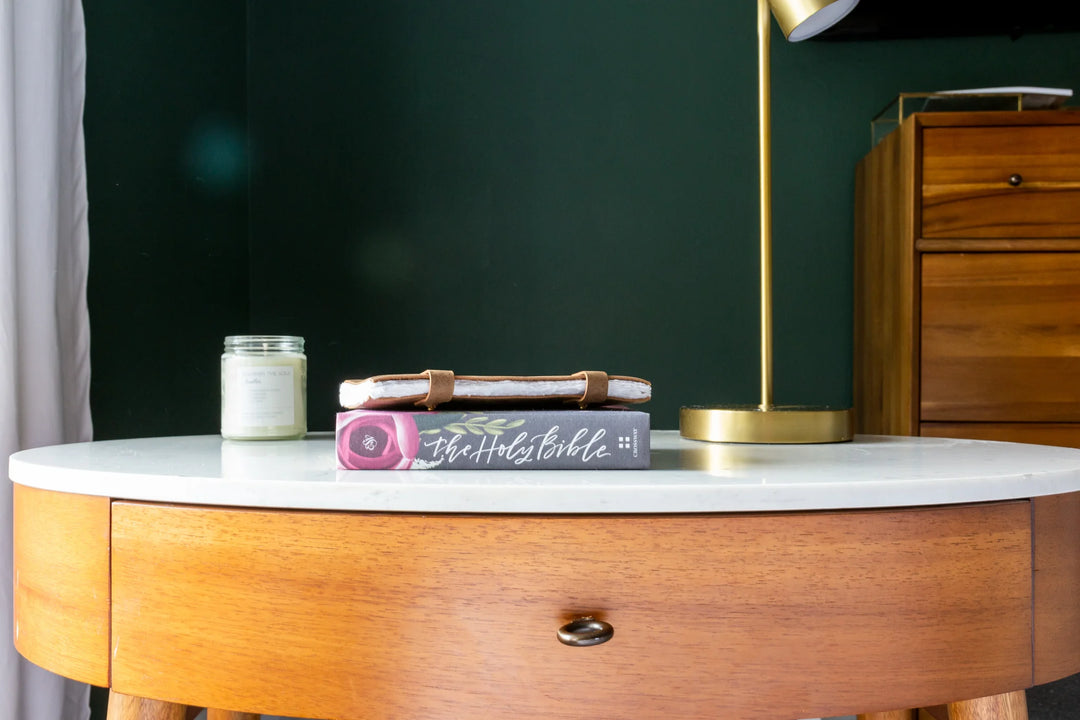 Wooden table with a white marble top, featuring a Bible and a journal tied with leather on it, beside a candle.