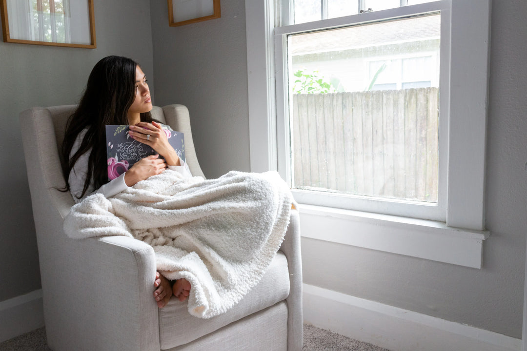 A woman sits in a light gray armchair, hugging a book, wrapped in a soft white blanket, gazing out the window.
