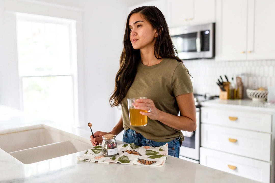 Young woman in a green shirt holds a glass of amber liquid near a jar of herbs and a patterned towel on a kitchen counter. What to Send When You Don't Know What to Say: A Sympathy Gift Guide.