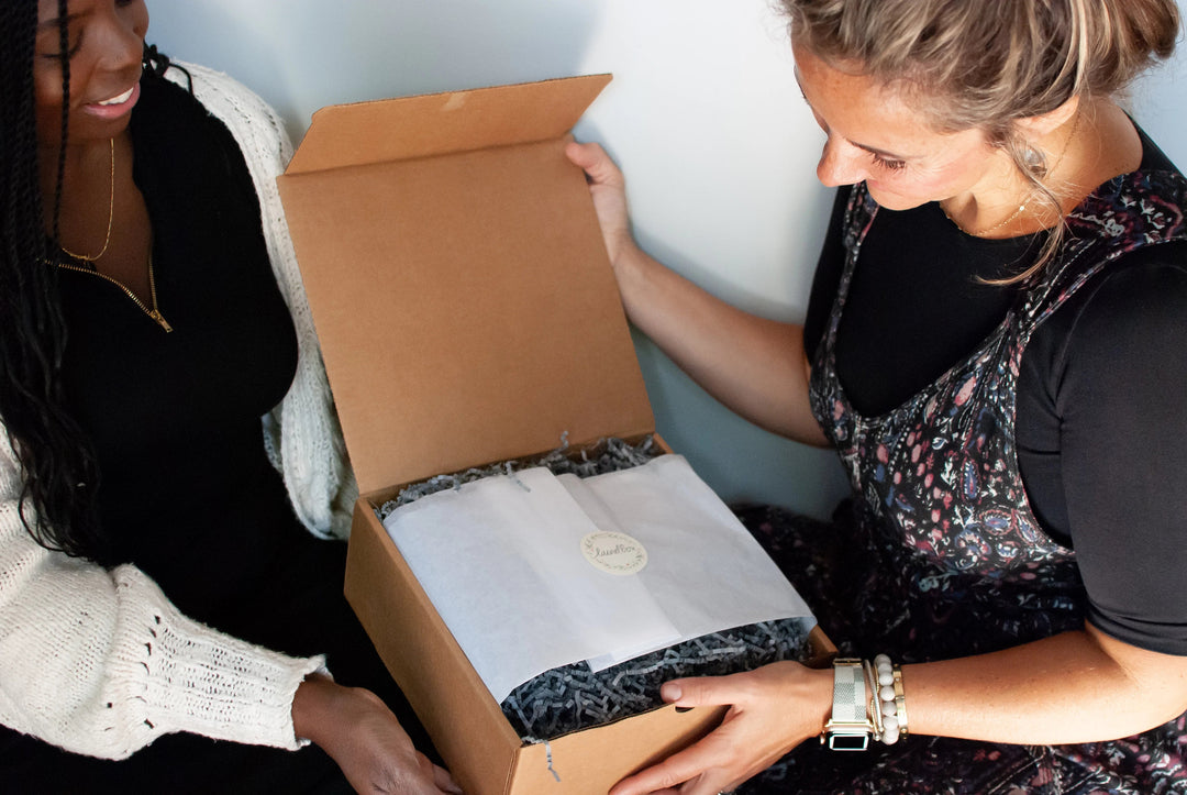 Two women opening a condolence gift box filled with black packing material and wrapped in white tissue.