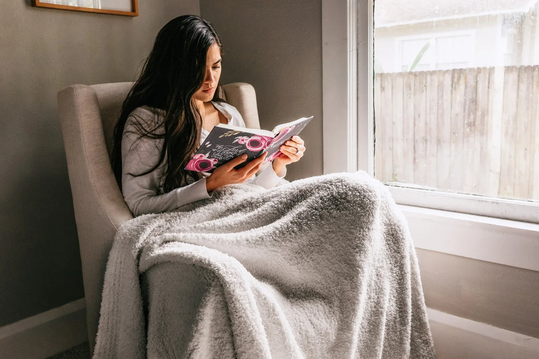 Woman with long hair sitting in a light gray armchair reading a book, wrapped in a soft, fluffy gray blanket, by a window.