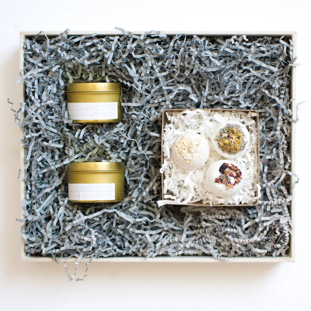 Gifts For All the Ladies on Your List: gold candle tins and bath bombs in a gift box with gray paper filler.