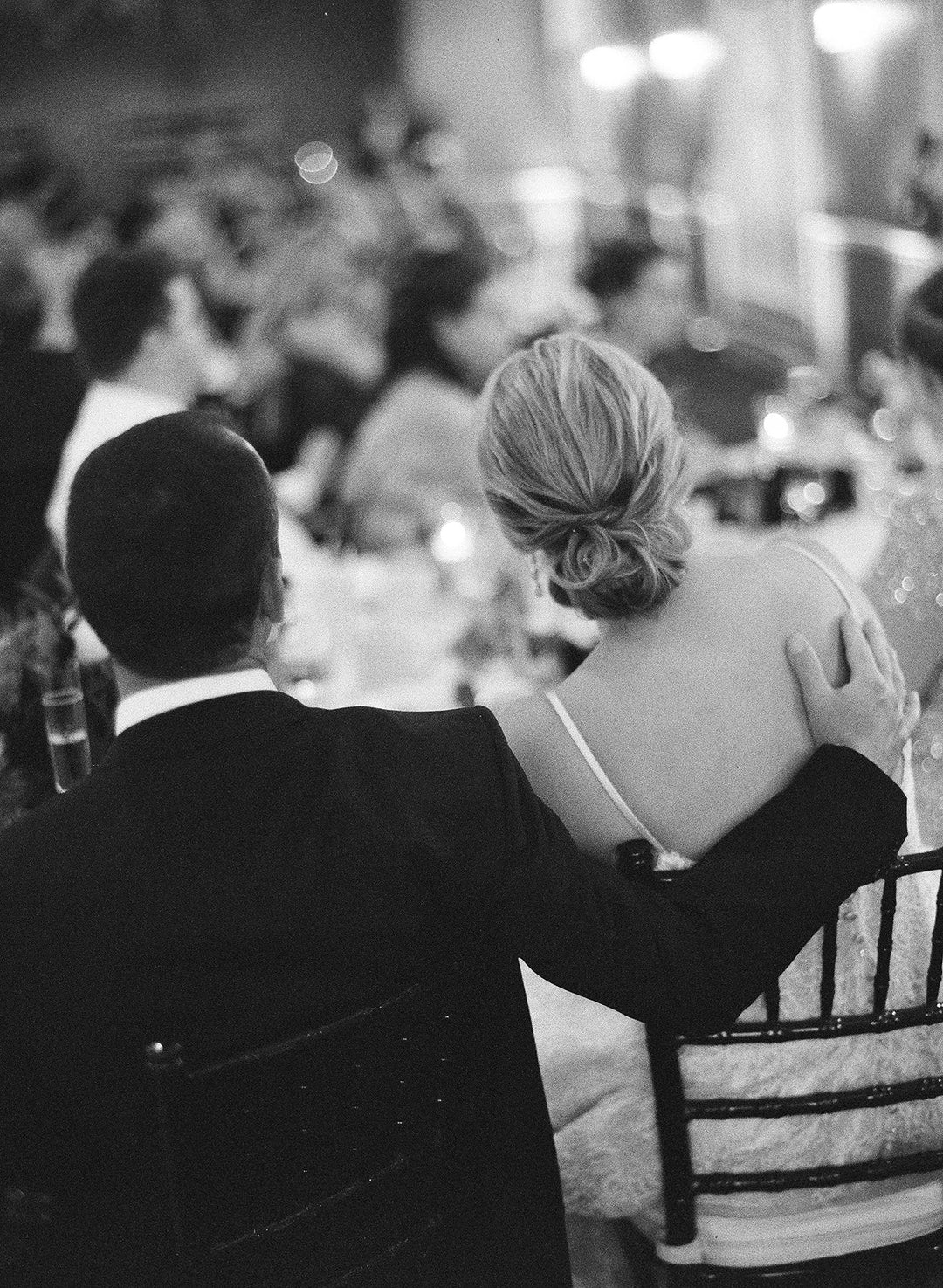 Couple in formal attire sitting together, woman in a white gown with a low back, man in a black suit, at a wedding reception. Memorial Candles As Wedding Gifts To Honor The Memory of a Loved One.