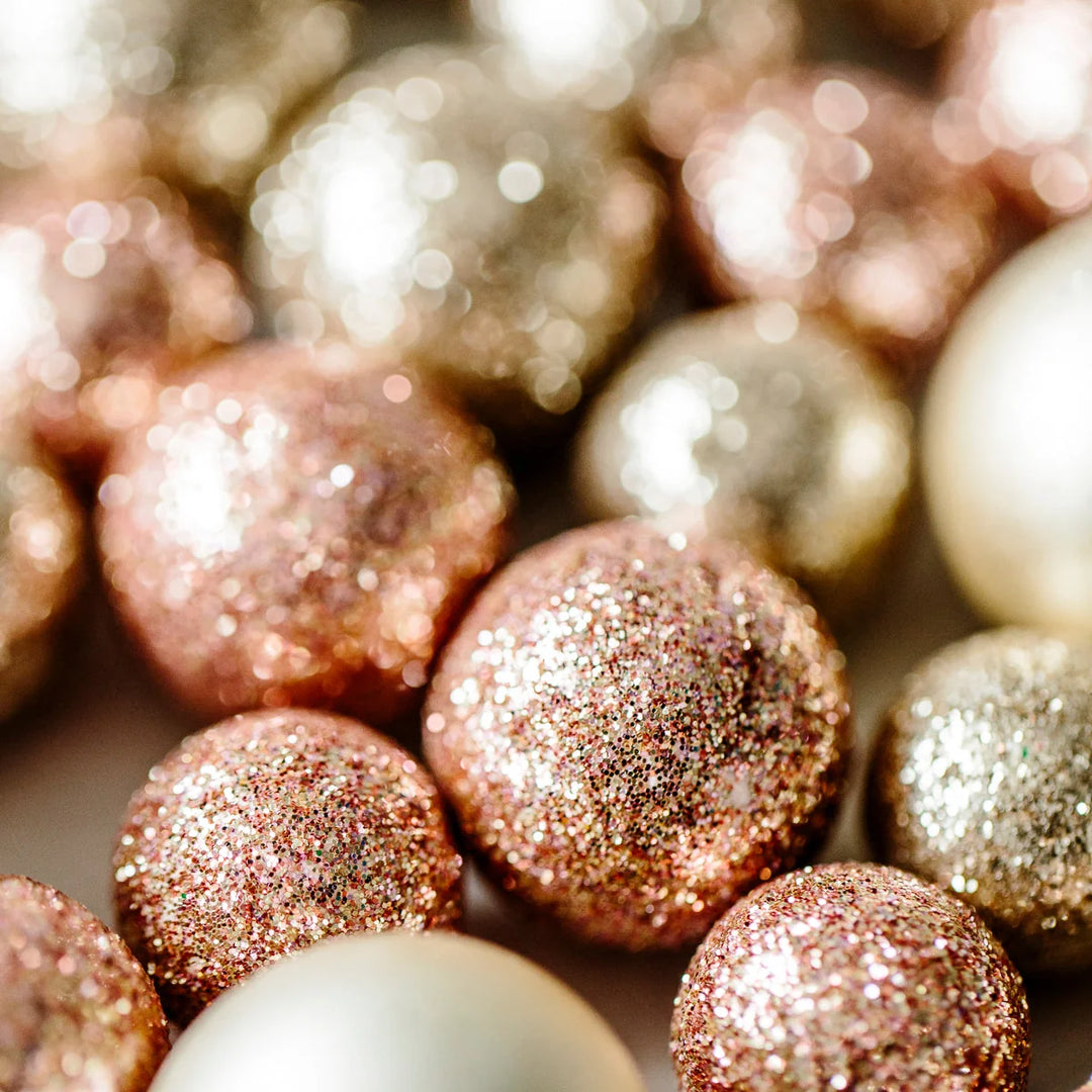 Shiny round ornaments in rose gold, gold, and silver glitter, varying in size, scattered together for A Holiday Gift-Giving Guide For Those Who Are Grieving.