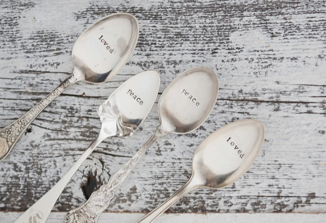 Four vintage silver spoons with engraved words "loved" and "peace" on the bowls, resting on a rustic wooden surface.