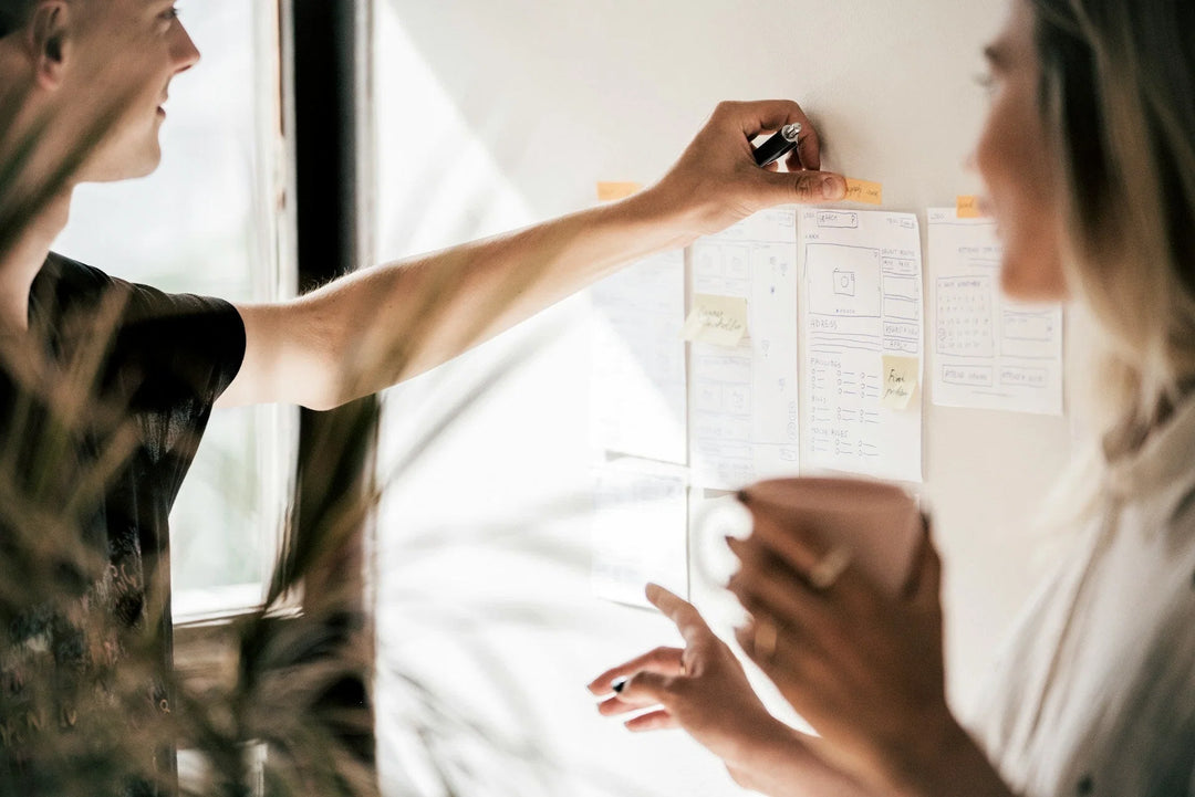 Two individuals discussing notes and sketches on a wall, one holding a cup, in a workspace setting. The Complete 2026 Guide to Thoughtful Workplace Sympathy Gifts.