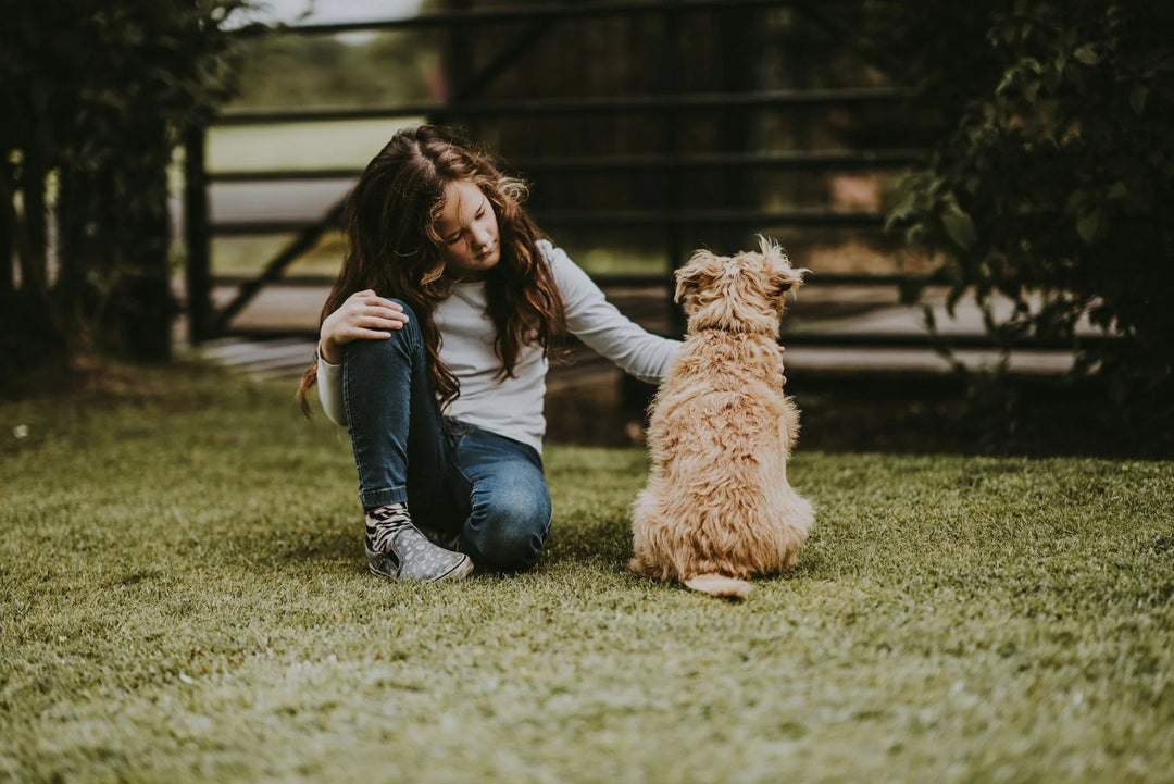 A girl in a white shirt and jeans sits on grass, reaching to pet a small, fluffy, tan dog, surrounded by greenery. The Best Memorial Gifts for Someone Who Lost a Pet.