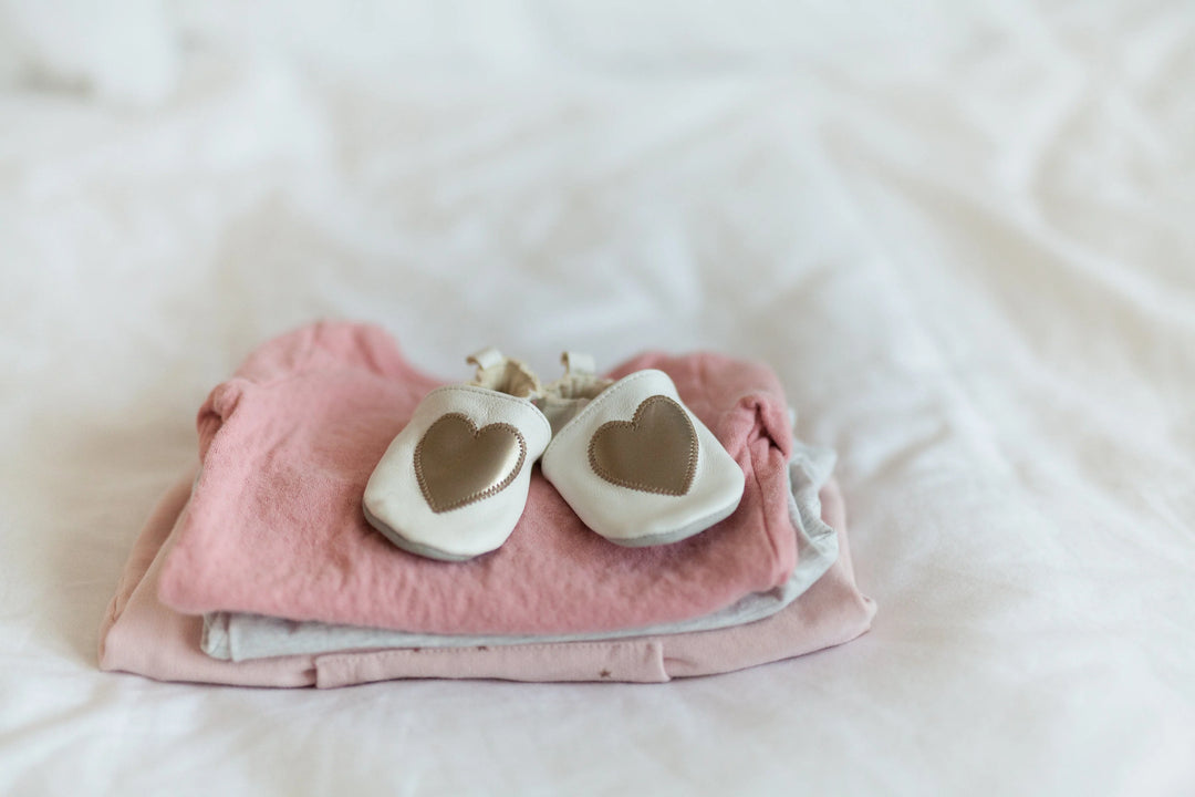 Light pink and gray infant clothing stacked with white shoes featuring a gold heart, related to 25 Meaningful Ways to Support Someone After Baby Loss in 2026.