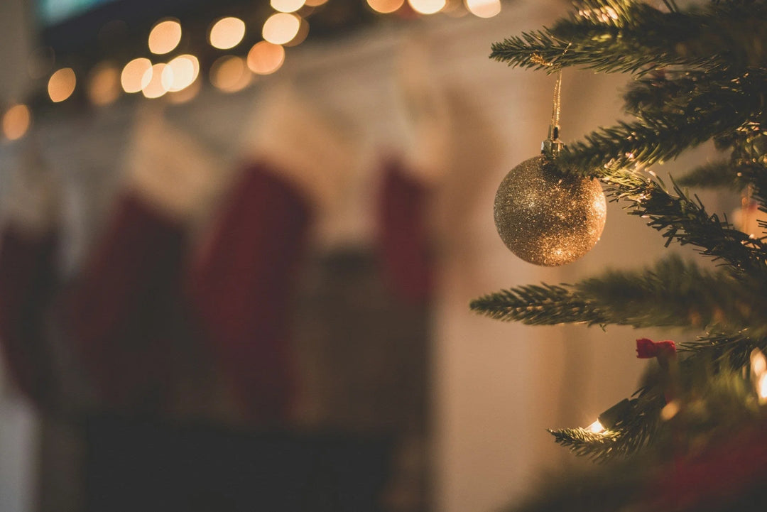 Gold glitter ornament hanging from a Christmas tree branch with softly blurred stockings in the background. Memorial Gifts for the First Christmas Without a Loved One (2026)