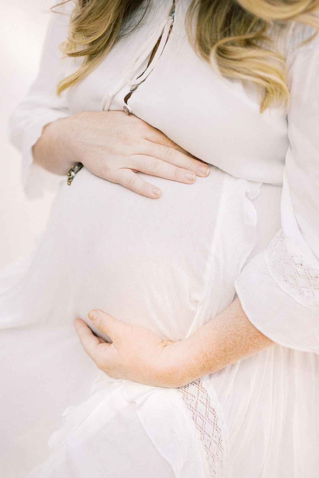 Hands resting on a pregnant belly, wearing a white flowing dress with lace details, focus on the gentle curve.