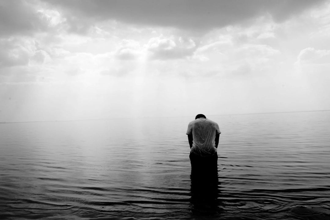 Man in wet gray shirt standing in shallow water, head bowed, with rippling water reflecting a cloudy sky. 15 Thoughtful Sympathy Gifts for Men Grieving a Loved One.