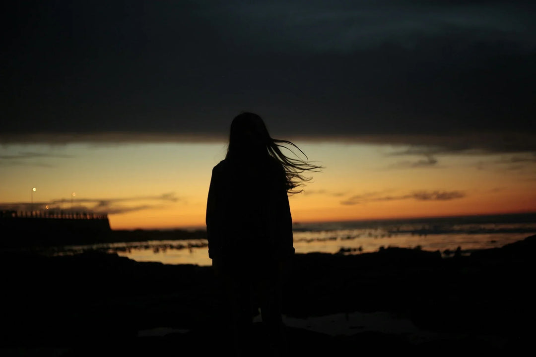 Silhouette of a woman with long hair against a sunset sky, standing by the water. Focus keyword: Navigating Grief Alone: Strategies for Self-Healing.