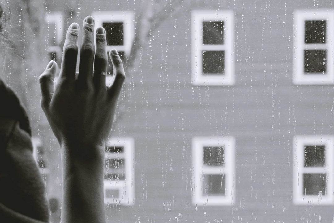 A hand with a ring pressed against a rainy window, with blurred window panes visible in the background. How to Support a Loved One Grieving a Loss From Suicide.