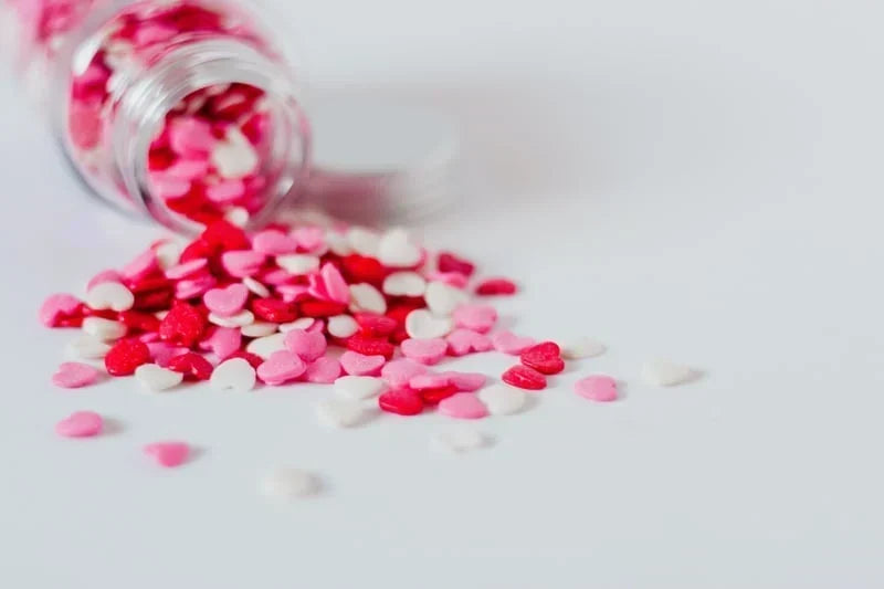 Colorful heart-shaped sprinkles in pink, red, and white spilling from a clear glass jar on a white surface.
