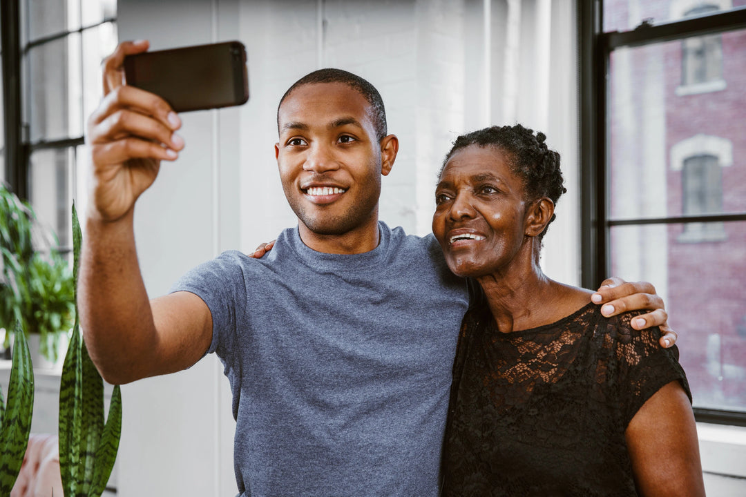 A young man in a blue shirt takes a selfie with an older woman in a black lace top, both smiling. 24 Thoughtful Sympathy Gifts for the Loss of a Grandparent.