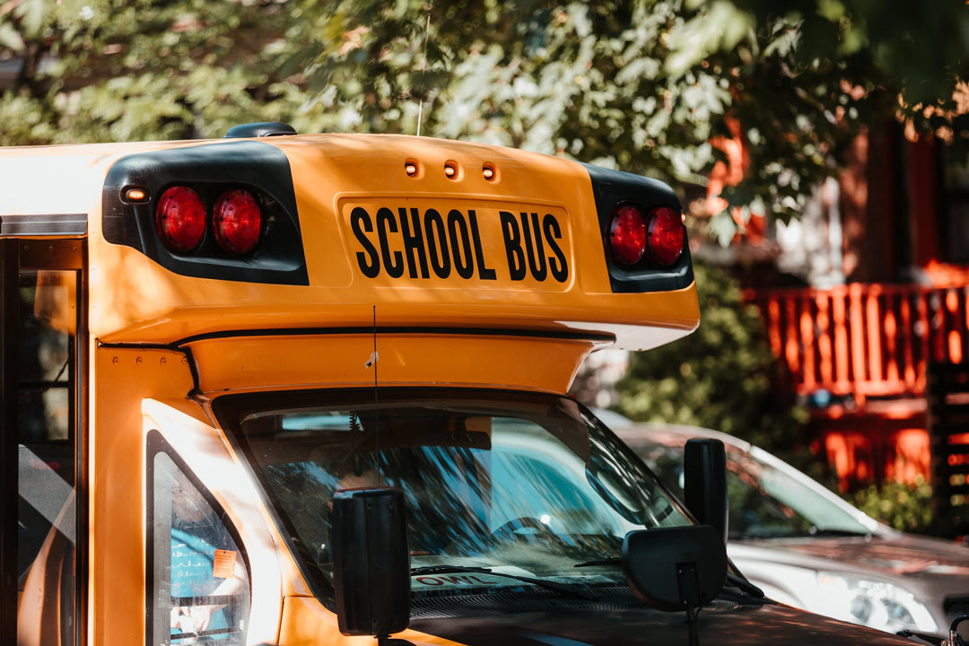 Yellow school bus with "SCHOOL BUS" text on the front, visible lights, and side mirrors, surrounded by green trees.